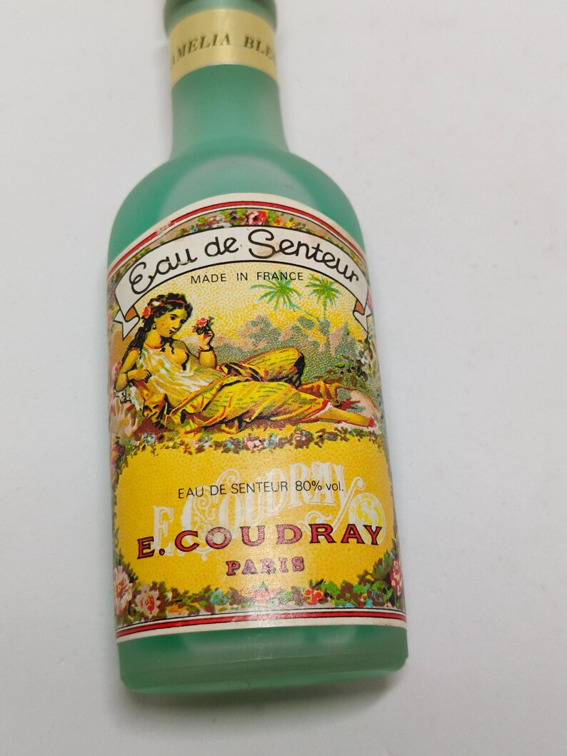 Puede incluir: Una botella de perfume vintage de vidrio verde claro con una etiqueta amarilla que presenta una mujer y dise&ntilde;os florales. La etiqueta dice "Eau de Senteur" y "E. Coudray, Paris". La botella tiene una tapa verde y probablemente es de Francia.