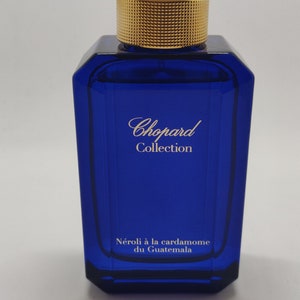 Könnte beinhalten: Eine blaue Glasflasche des Chopard Collection Néroli à la cardamome du Guatemala Parfüms. Die Flasche hat einen goldenen Verschluss mit dem Chopard Logo.
