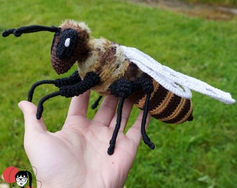 May include: A crocheted bee toy with brown and yellow stripes, black legs, and white wings. The bee is sitting on a person's hand.