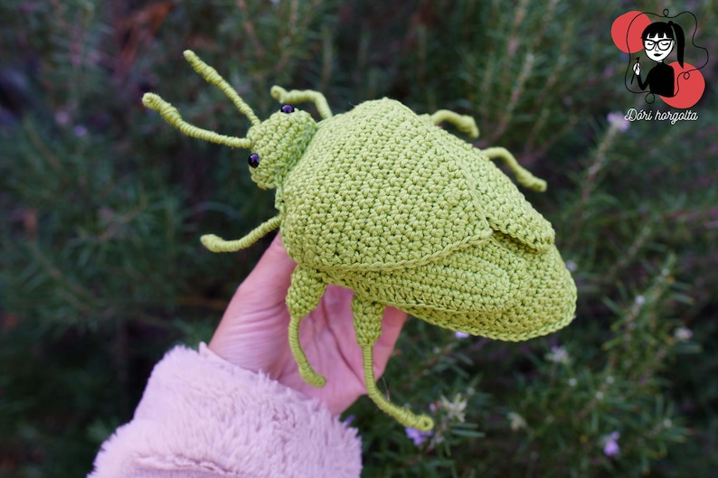 May include: A handmade, light green crochet insect toy with black eyes and long antennae. The insect is held against a blurred green background. The toy is a unique, whimsical creation.