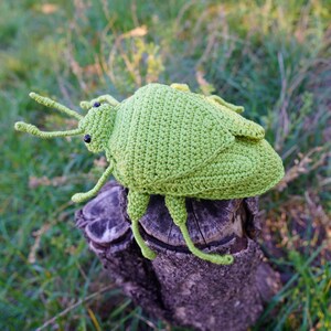 May include: A handmade, light green crochet insect toy with black eyes and long antennae. The insect is perched on a weathered wooden stump, set against a blurred background of green grass and foliage. The toy is a whimsical, handcrafted item.