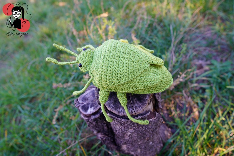 May include: A handmade, light green crochet insect toy with black eyes and long antennae. The insect is perched on a weathered wooden stump, set against a blurred background of green grass and foliage. The toy is a whimsical, handcrafted item.