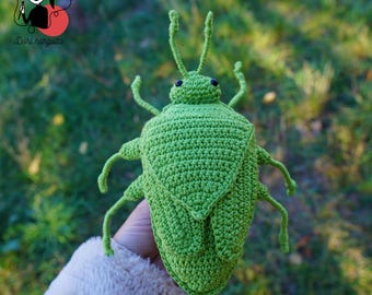 May include: A handmade, green crochet insect with detailed antennae, legs, and body segments. The bug has small, black eyes and is held against a blurred green background. The image includes the text "Dóri horgolta".