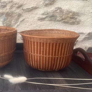 Peut inclure: Deux paniers en osier tressés, l'un plus grand que l'autre, sont posés sur une surface en bois foncé. Le plus grand panier est brun et de forme arrondie. Le plus petit panier est également brun et de forme plus cylindrique.