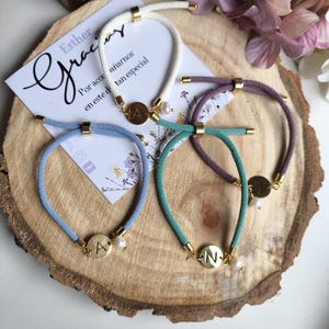 Puede incluir: Conjunto de pulseras de cuerda en varios colores, incluyendo blanco, azul, turquesa y morado. Cada pulsera tiene detalles metálicos dorados y un dije con una inicial. Algunas pulseras también incluyen una pequeña perla. Hay una tarjeta con texto manuscrito.