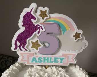 May include: A white cake with white frosting and a unicorn cake topper. The topper features a purple glitter unicorn, a rainbow, a cloud, stars, and the number 5. The banner says "ASHLEY".