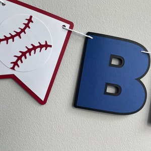 May include: A baseball-themed banner with the letters "BIG" in blue with black outlines. The letter "B" is shown in the image. The banner is strung with white string and has a red and white pennant with a baseball graphic.