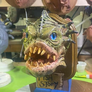 May include: A detailed sculpture of a creature's head with green and tan skin, sharp teeth, and blue eyes. The sculpture is mounted on a wooden plaque with the words "FISH STORY" on a small sign.