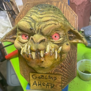 May include: A goblin head sculpture with red eyes and sharp teeth, mounted on a wooden plaque. A small sign on the plaque reads "Goblins Ahead". The goblin is painted in shades of green and brown.