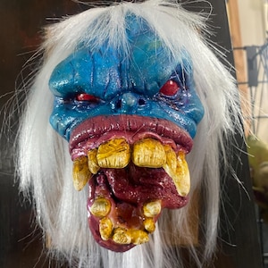 May include: A grotesque mask with a blue face, red eyes, and a wide, open mouth revealing yellow teeth. The mask has white, shaggy hair and a textured appearance, creating a frightening aesthetic.