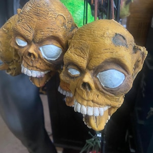 May include: Close-up of several zombie skull ornaments. The skulls are painted in shades of yellow and brown, with white eyes and exposed teeth. The tongues are visible. The ornaments are hanging from brown cords, suggesting they are for decoration.