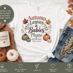 May include: A white short-sleeve t-shirt featuring a graphic print with the text Autumn Leaves & Babies Please, surrounded by illustrations of autumn leaves, sprigs, and four smiling pumpkins in shades of green and orange, with a small heart icon.