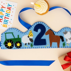 Puede incluir: Una corona de cumpleaños de fieltro azul con un diseño de tractor, oveja, caballo y vaca, con el número 2. Incluye una cinta azul, una magdalena con una vela encendida y una tarjeta que dice "Happy Birthday". También hay un regalo envuelto con un lazo rojo.