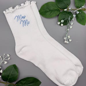 May include: A pair of white ankle socks featuring a ruffled top edge and the text "Mrs Me" embroidered in light blue script. The socks are made of a ribbed knit fabric and are displayed alongside green leaves and small white floral buds.