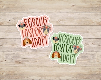 May include: Two stickers with a green and pink background. Both stickers feature the text "Rescue Foster Adopt" with a stylized font and three cartoon dogs with flowers on their heads.