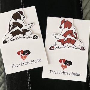 May include: Two white cards with brown and red illustrations of Brittany Spaniel dogs in a sitting position. The cards have the text "Three Britts Studio" printed on them.
