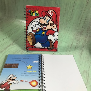 Può includere: Un taccuino a spirale con una copertina rossa raffigurante il personaggio di Super Mario. La copertina mostra le parole "Super Mario" e un'immagine di Mario. Le pagine aperte mostrano illustrazioni e testo sulla Super Stella.