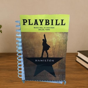 Puede incluir: Un cuaderno de espiral con una portada de Playbill de Hamilton. La portada muestra el texto PLAYBILL, MUSIC HALL AT FAIR PARK, DALLAS, TEXAS y HAMILTON, con la silueta de una persona sobre una gran estrella negra contra un fondo dorado.