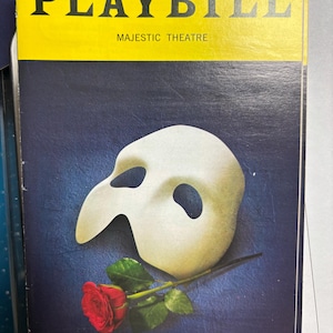 Puede incluir: Una portada de Playbill con el texto "PLAYBILL" en amarillo sobre "MAJESTIC THEATRE". La portada tiene un fondo azul oscuro, una máscara blanca y una rosa roja.