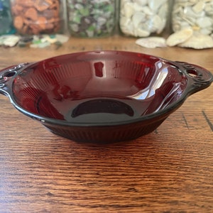 May include: A deep red glass bowl with two handles. The bowl has a ribbed design and is made of clear glass.