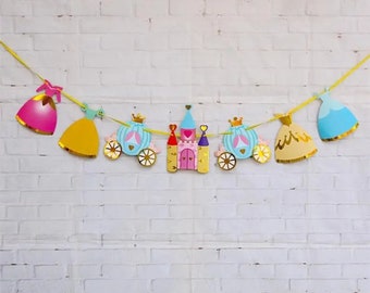 Op de afbeelding: Een prinsessen-thema feestbanner met een gele string, met een roze, gele en blauwe jurk, een blauwe koets, een roze kasteel en een gouden kroon.