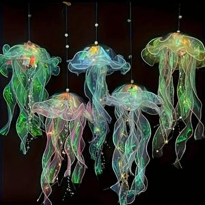 May include: Five hanging jellyfish decorations with translucent, flowing forms. The jellyfish are primarily green with hints of pink and blue, and have long, ribbon-like tentacles. Small, clear beads are suspended from the top, and the background is black.