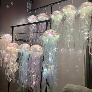 May include: A collection of iridescent jellyfish-shaped hanging lights. Each light features a ruffled, translucent cap and flowing, ribbon-like tentacles. The lights are suspended from a black metal frame, creating a whimsical and ethereal display.