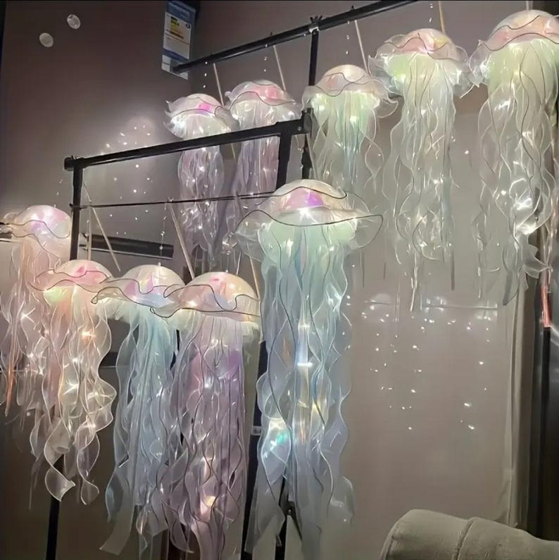 May include: A collection of iridescent jellyfish-shaped hanging lights. Each light features a ruffled, translucent cap and flowing, ribbon-like tentacles. The lights are suspended from a black metal frame, creating a whimsical and ethereal display.