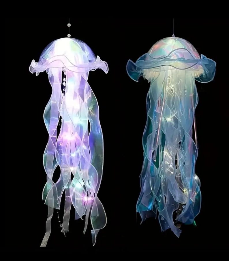 May include: Two hanging jellyfish decorations with iridescent, flowing tentacles and illuminated bodies. The jellyfish have a translucent, ruffled cap and are suspended from thin wires against a black background. The lights inside the jellyfish glow with a soft, colorful light.