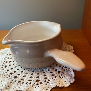 May include: A light pink ceramic sauce pot with a white interior and a handle. The pot is sitting on a white crocheted doily.