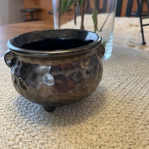 Puede incluir: Un pequeño macetero de cerámica negro con forma de caldero con tres patas. El macetero tiene un acabado de metal martillado.