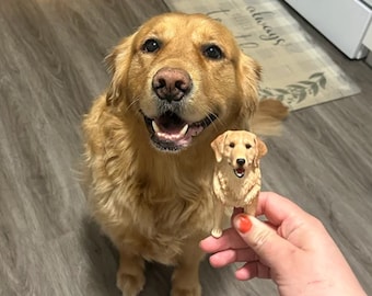 May include: A golden retriever dog looks up at a 3D printed figurine of a golden retriever dog. The figurine is being held in a person's hand.