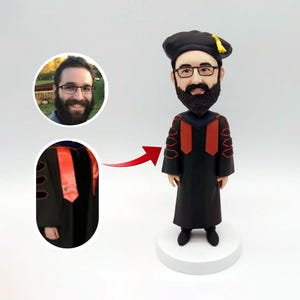 Könnte beinhalten: Eine maßgefertigte Figur einer Person, die eine schwarze Graduierungsrobe mit roten Akzenten und eine schwarze Kappe mit einer goldenen Quaste trägt. Die Figur steht auf einem weißen Sockel.