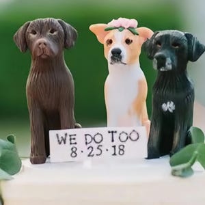 May include: Three dog figurines, a brown labrador, a white and tan chihuahua wearing a flower crown, and a black labrador, are arranged on a white cake stand. The cake stand has a sign that reads "We Do Too 8.25.18".
