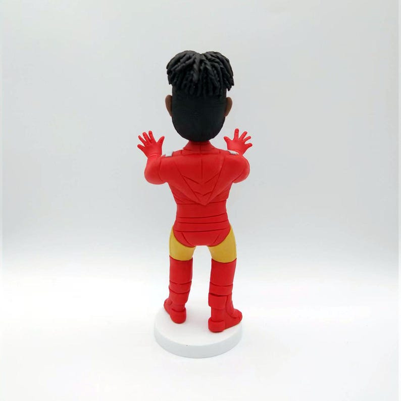 May include: A red and yellow figurine of a person wearing a superhero costume. The figurine is standing with its arms raised and is facing away from the camera.