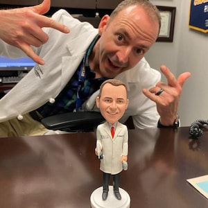 May include: A white lab coat bobblehead figurine of a doctor holding a stethoscope and a syringe. The figurine is standing on a white base. The bobblehead is in front of a person wearing a white lab coat and making a rock and roll hand gesture.