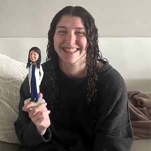 Puede incluir: Una persona sosteniendo una pequeña figura de una persona que lleva una bata de laboratorio blanca y pantalones azules. La figura está de pie sobre una base blanca.