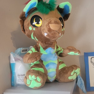 May include: A brown plush toy with green and blue accents. The toy has a long tail, large eyes, and a toothy grin. It is sitting on a white dresser.