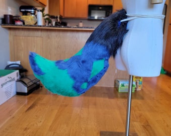 May include: A blue, green, and black faux fur tail attached to a white mannequin. The tail is a vibrant mix of colors, with a dark blue tip and a pattern of green and blue on the body. The tail is attached with a white cord.