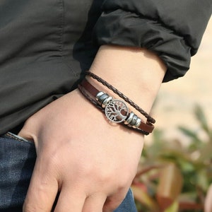 Puede incluir: Un brazalete de cuero marrón con un dije de árbol de la vida plateado y cuentas plateadas. El brazalete está trenzado y tiene un diseño en capas.