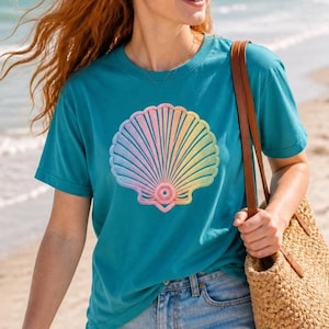May include: Teal t-shirt featuring a large, embroidered seashell design with pastel rainbow colors. The shirt is paired with blue jeans and a woven tote bag with a brown leather strap. The background shows a beach scene.