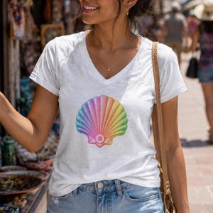 Może przedstawiać: Biały t-shirt z krótkim rękawem i dekoltem w serek, ozdobiony centralną grafiką muszli z żywym, tęczowym gradientem. Wzór muszli zawiera przejścia kolorystyczne obejmujące odcienie niebieskiego, fioletowego, różowego, pomarańczowego, żółtego i zielonego.
