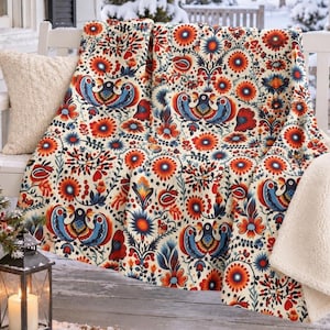 May include: A throw blanket featuring a cream background with a repeating folk-art pattern of stylised blue birds, vibrant orange and red flowers, and green foliage. The blanket has a soft, white sherpa-style fleece lining on the reverse side.