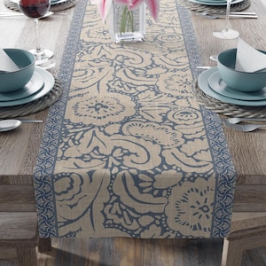 Op de afbeelding: Een rechthoekige tafelloper met een beige basis, een donkerblauw bloemmotief en een decoratieve rand. De loper ligt in het midden van een houten tafel, geflankeerd door blauwe kommen, witte servetten, wijnglazen en bestek op gevlochten placemats.