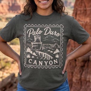 May include: A dark olive green short-sleeve t-shirt featuring a cream-colored graphic print shaped like a postage stamp. The design depicts a desert landscape with rock formations and the text Palo Duro CANYON in a stylized script and serif font.