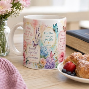 May include: A white ceramic mug with a floral design and Spanish text, including butterflies. The mug has a curved handle and is on a wooden surface, next to a plate of pastries and fruit.