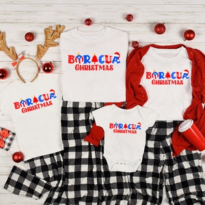 May include: A family matching Christmas pajama set with white shirts featuring the text "Boracua Christmas" in red, blue, and white. The shirts are worn by a child, an adult, and a baby. The adult is wearing red and black plaid pants.