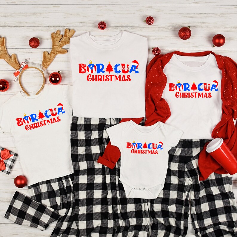 May include: A family matching Christmas pajama set with white shirts featuring the text "Boracua Christmas" in red, blue, and white. The shirts are worn by a child, an adult, and a baby. The adult is wearing red and black plaid pants.
