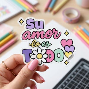 May include: A colorful sticker with the text "Su amor lo es TODO" in various colors, including purple, pink, and green. The design features a daisy and heart shapes. The sticker is held by a person with pink nail polish.