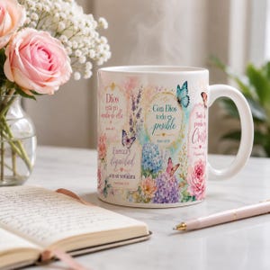 May include: White ceramic mug with floral and butterfly designs, and text in Spanish. The mug features pink, blue, and purple flowers, with butterflies. A book, pen, and flowers are in the background.